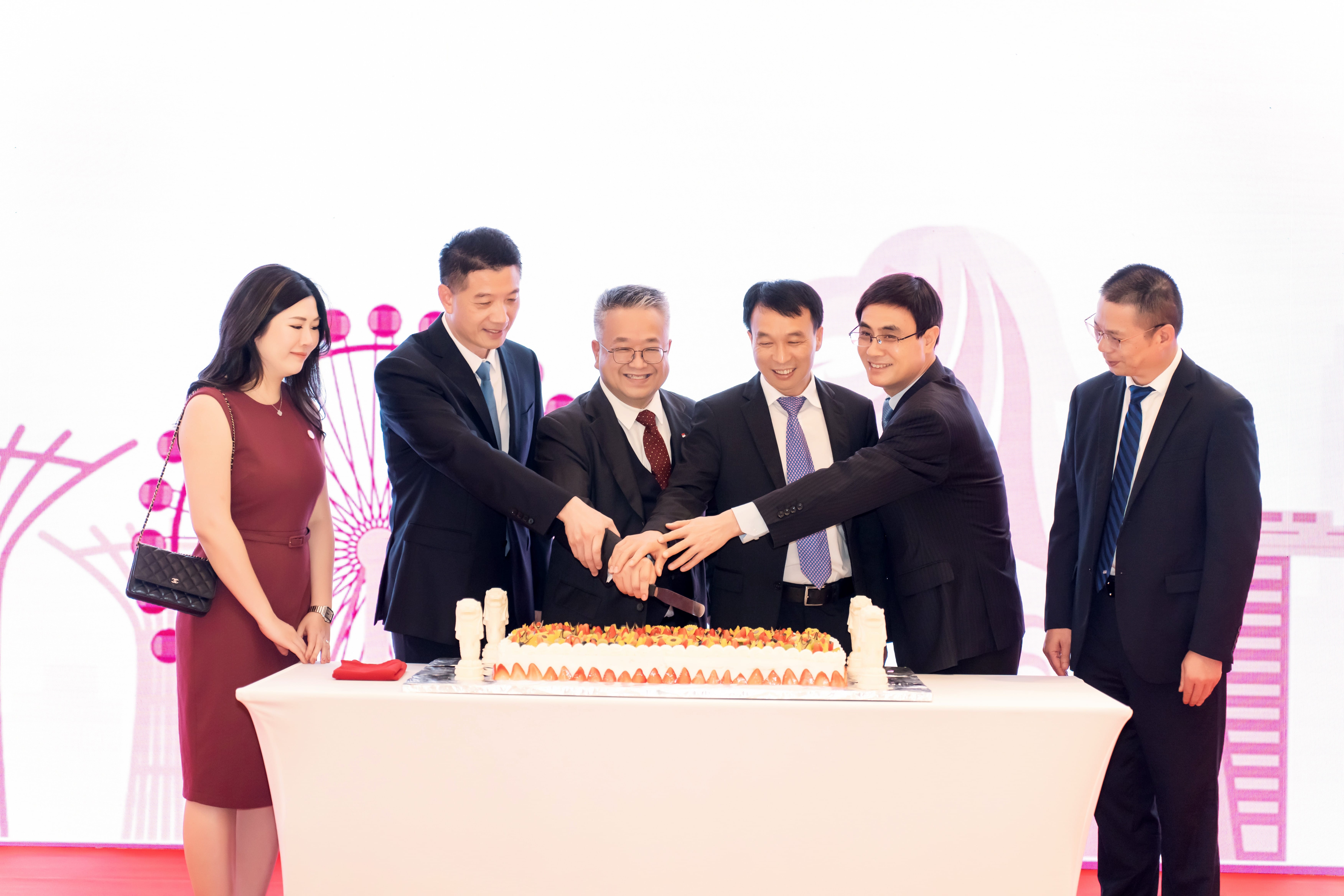 Group cutting fruit-topped cake on a white-clothed table. A woman in a red dress with Chanel bag stands nearby.
